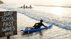 "A young surfer lying on a blue foam surfboard at the beach next to a wooden sign that says Surf School: First Waves Here."
