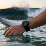 A close-up view of a surfer's wrist wearing a durable, waterproof fitness tracker while in the ocean, with a surfboard and ocean waves in the background at sunset. Perfect for tracking surf sessions. SurfingGears.com