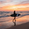 A woman carrying a black and white patterned longboard surfboard walks along a beach at sunset, with dramatic orange and pink hues reflected in the wet sand. SurfingGears.com