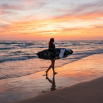 A woman carrying a black and white patterned longboard surfboard walks along a beach at sunset, with dramatic orange and pink hues reflected in the wet sand. SurfingGears.com